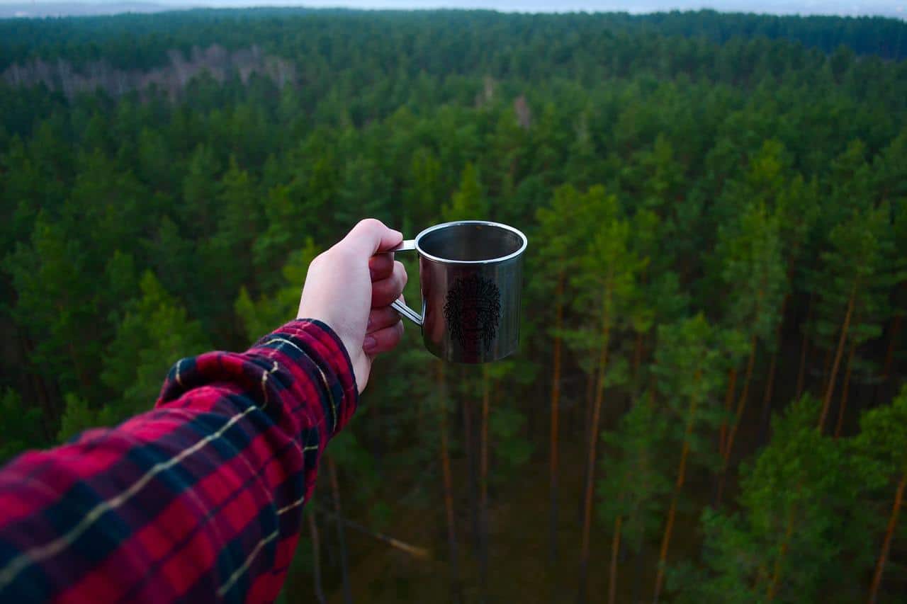 Kovový hrnek v ruce nad lesem. Turistika v přírodě, kempování, outdoorové aktivity. Dovolená v lese.