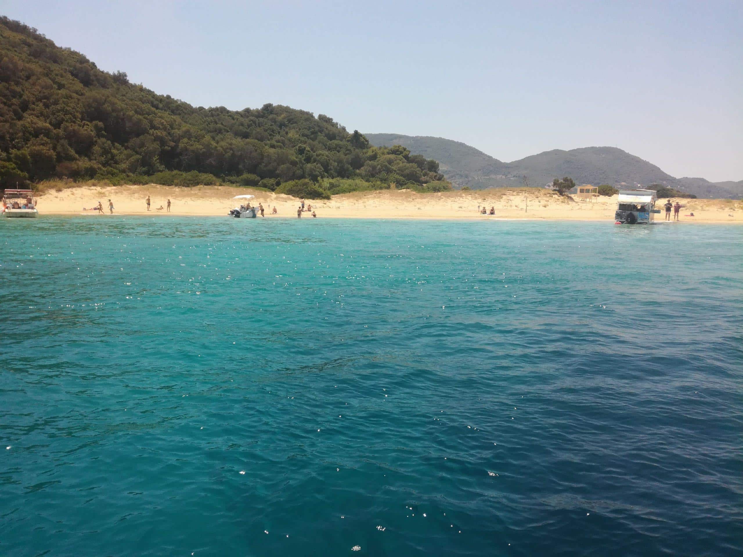 Pláž Laganas, Zakynthos. Hnízdiště želv, tyrkysové moře, písečná pláž s lidmi, zalesněný kopec. Dovolená v Řecku.