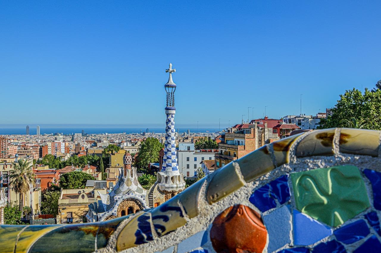Park Güell: Panorama Barcelony s mozaikou a věží. Gaudího architektura, slunce a výhled na město.