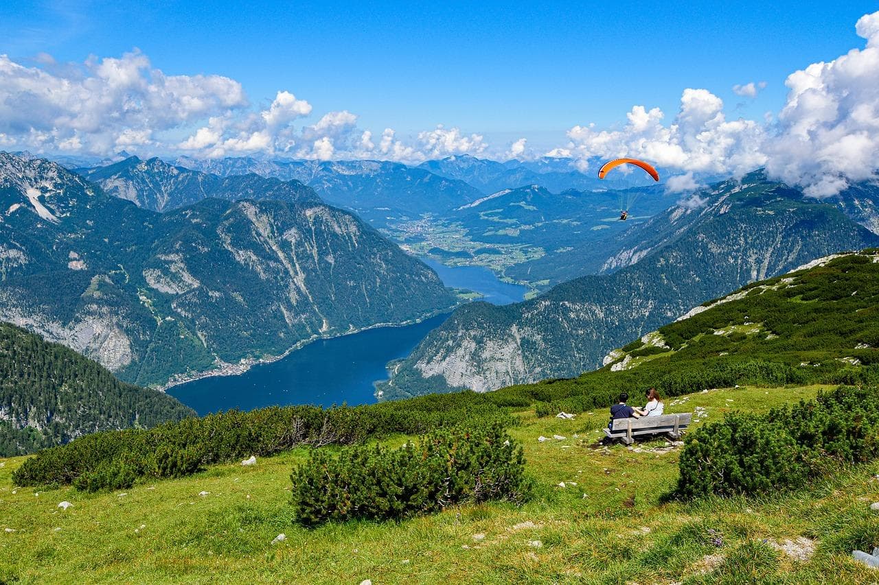 Rakouská jezera: Výhled na jezero z hor. Pár sedí na lavičce, paragliding.