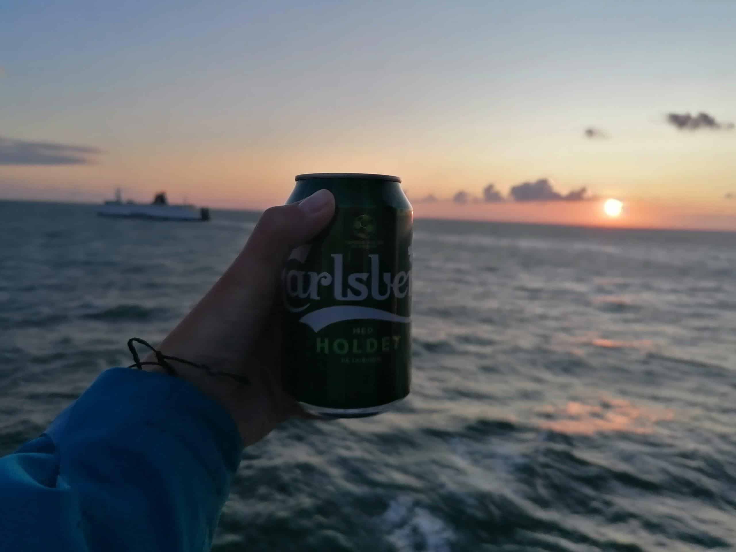 Pivo Carlsberg v ruce, západ slunce nad mořem. Pivovar Carlsberg.