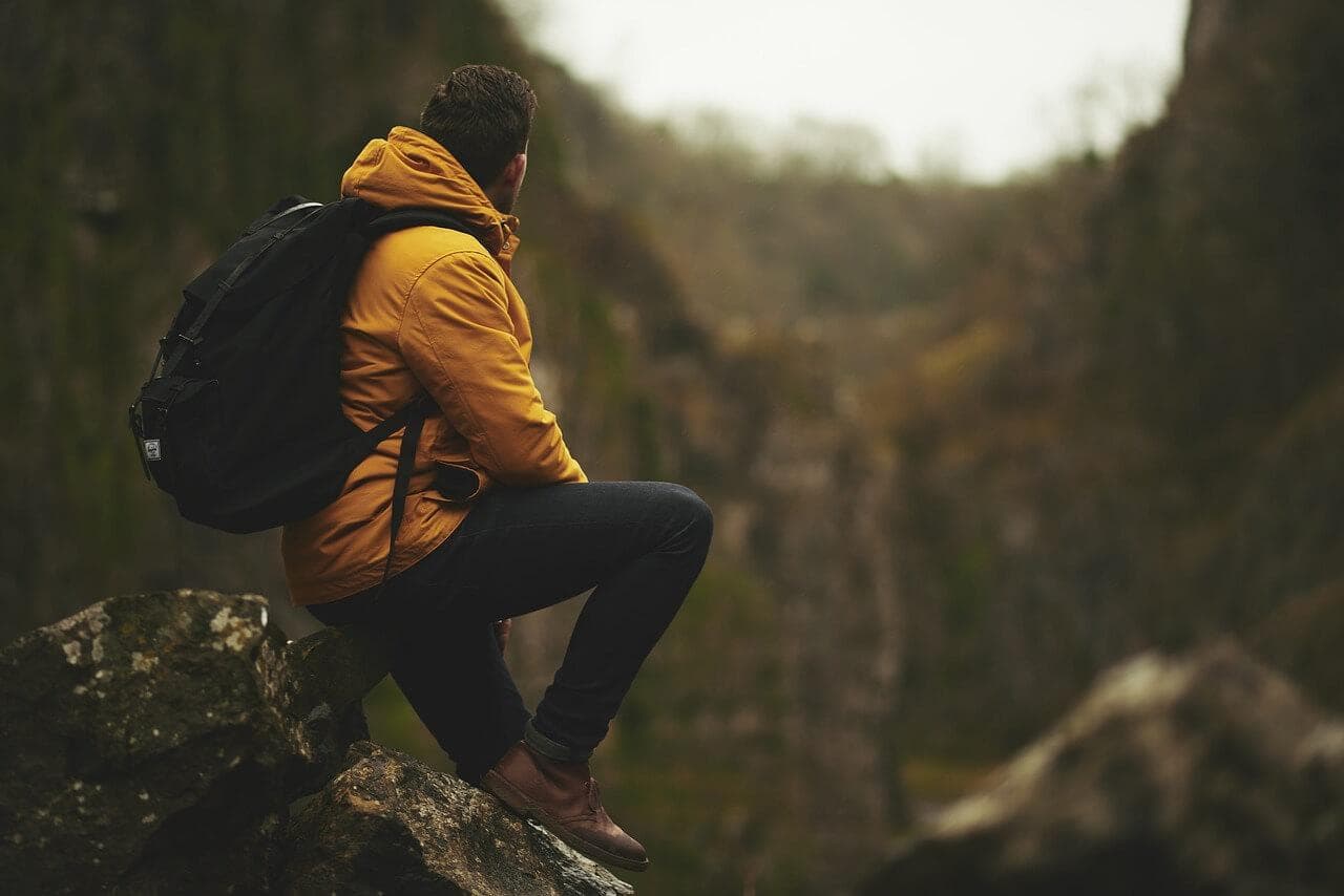Muž v outdoorovém oblečení s batohem sedí na skále a dívá se do krajiny.