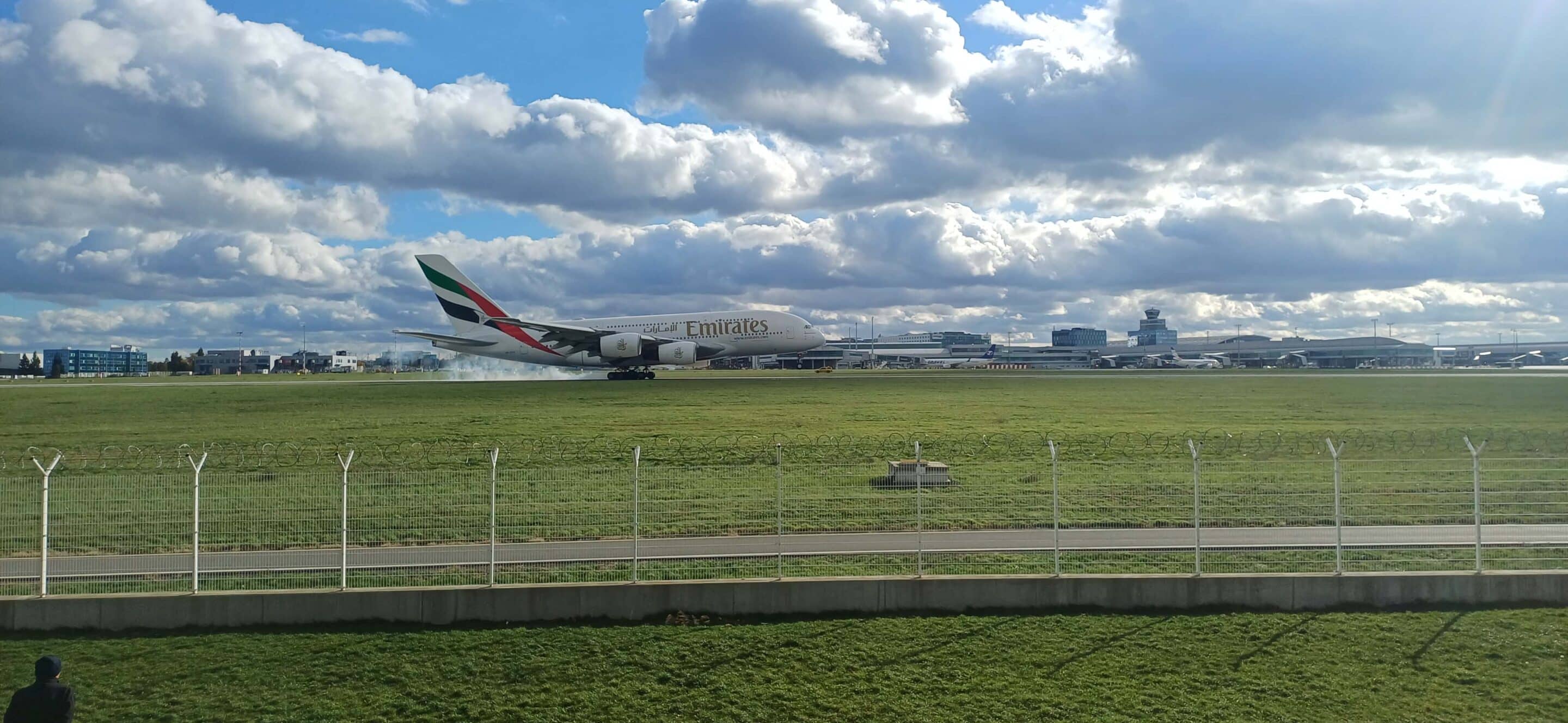 Letadlo Emirates A380 přistává na letišti. Zelená tráva, modrá obloha s mraky. Bezpečnost letecké dopravy a cestování.
