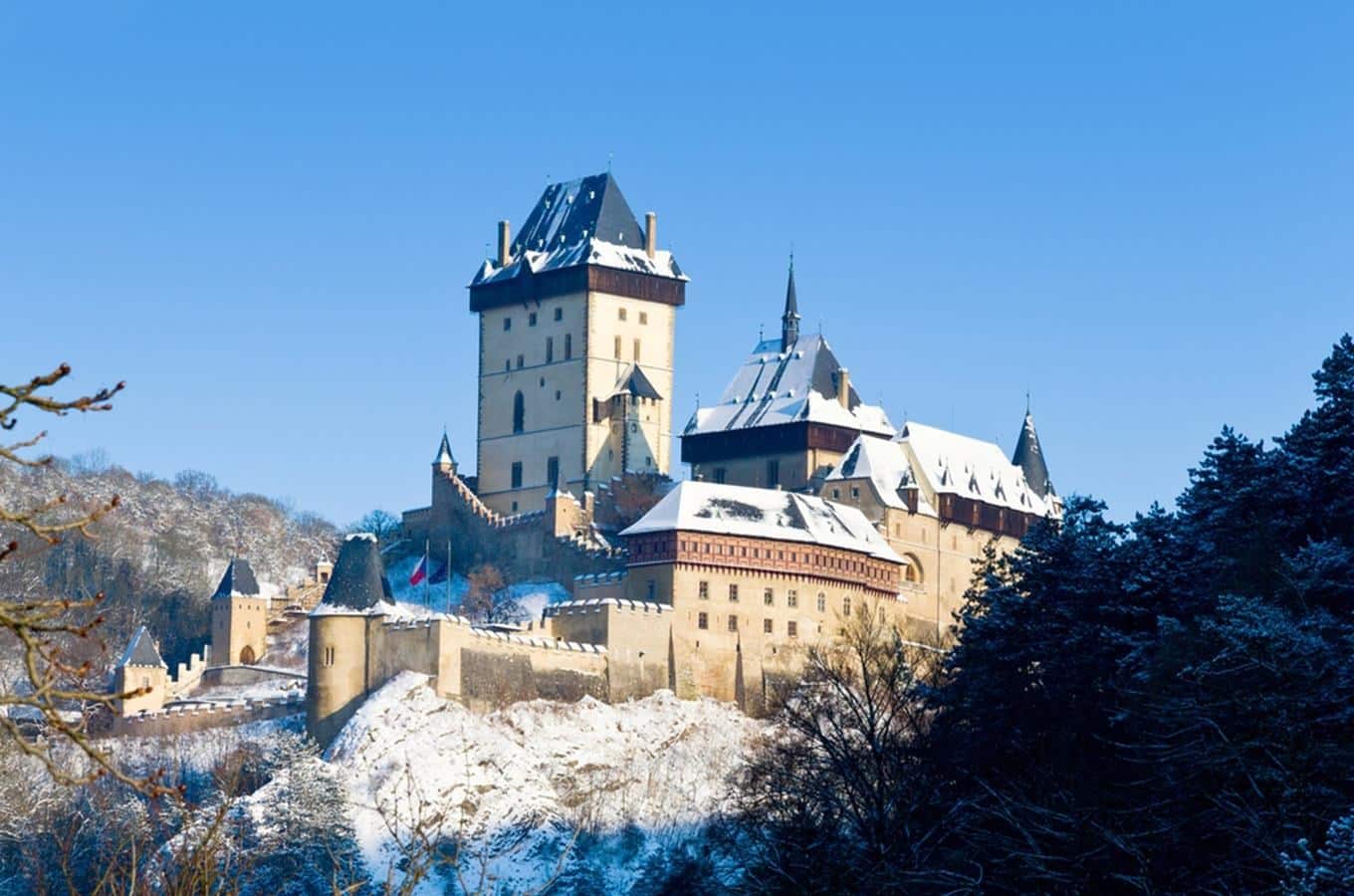 Hrad Karlštejn v zimě, zasněžená střecha, modrá obloha. Středověká architektura, česká památka, turistická destinace.
