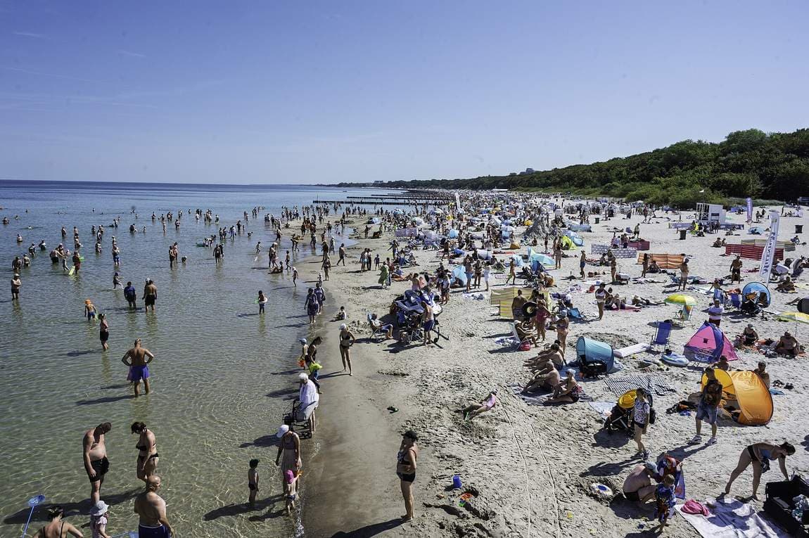 Široká písečná pláž v Kolobřehu s vlnolamy a davem lidí ve vodě za letního odpoledne