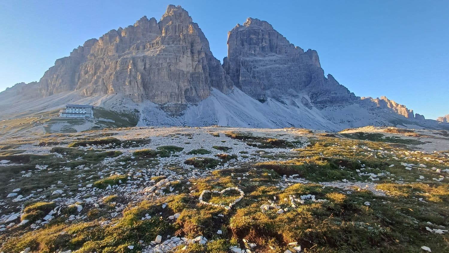 Srdce z kamenů v trávě, v pozadí vrcholy Tre Cime di Lavaredo a chata