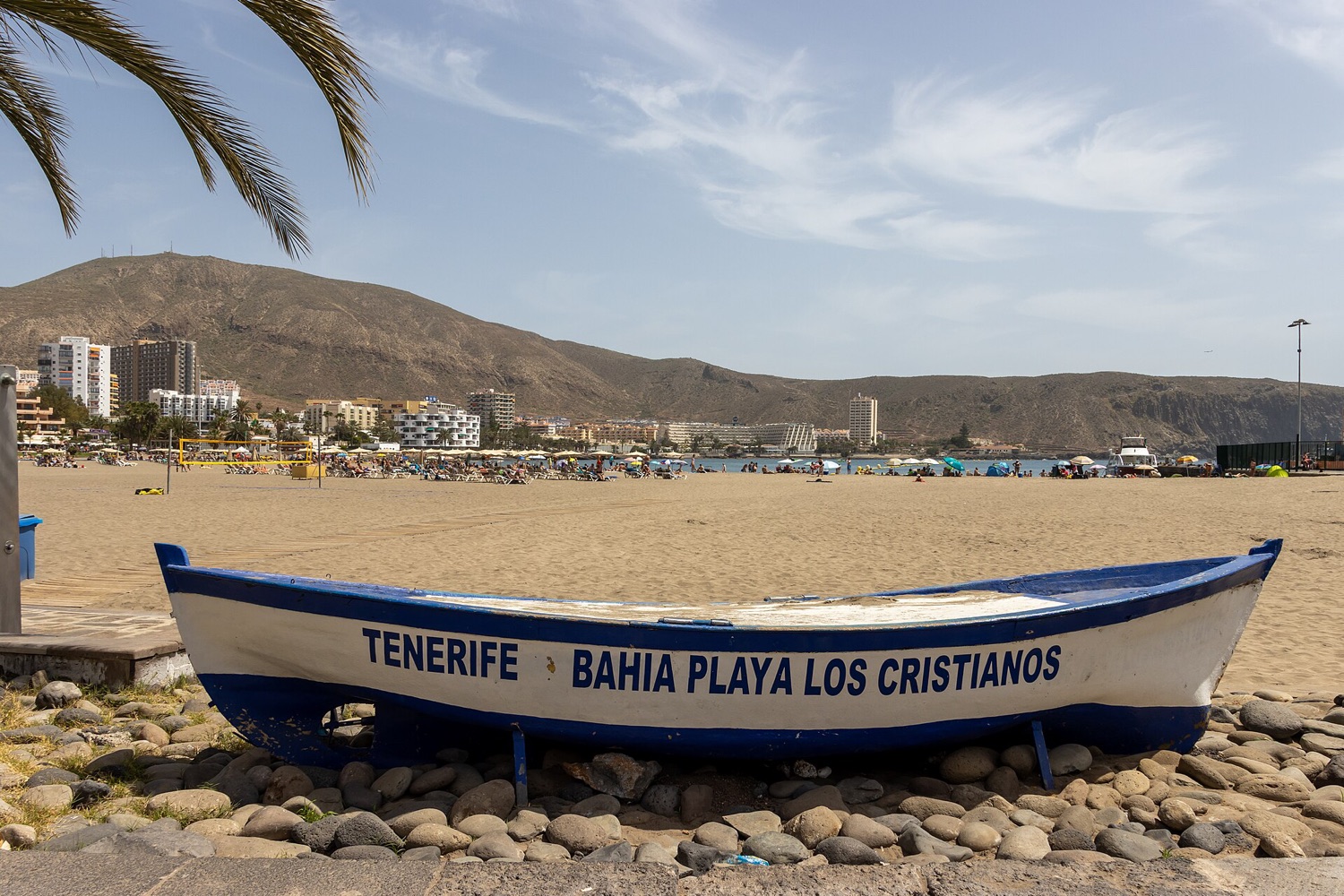 Pláž Los Cristianos na jihu Tenerife s loďkou a hotely pod suchými kopci