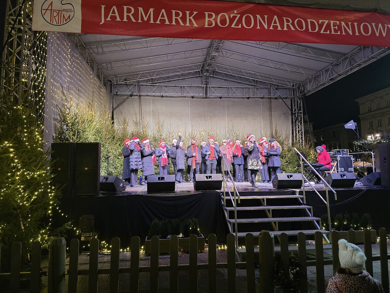 Sbor zpívá na pódiu vánočního trhu v Krakově pod nápisem Jarmark Bożonarodzeniowy