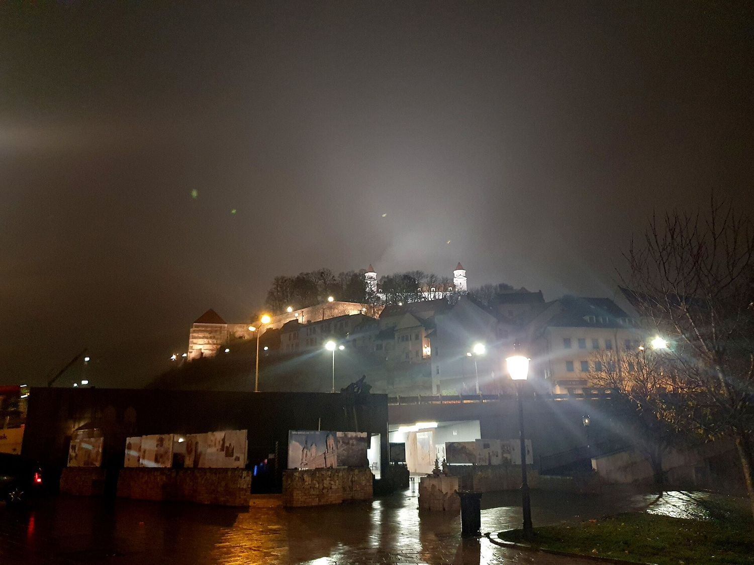 Bratislavský hrad na kopci v noci a v mlze, v popředí osvětlená ulice