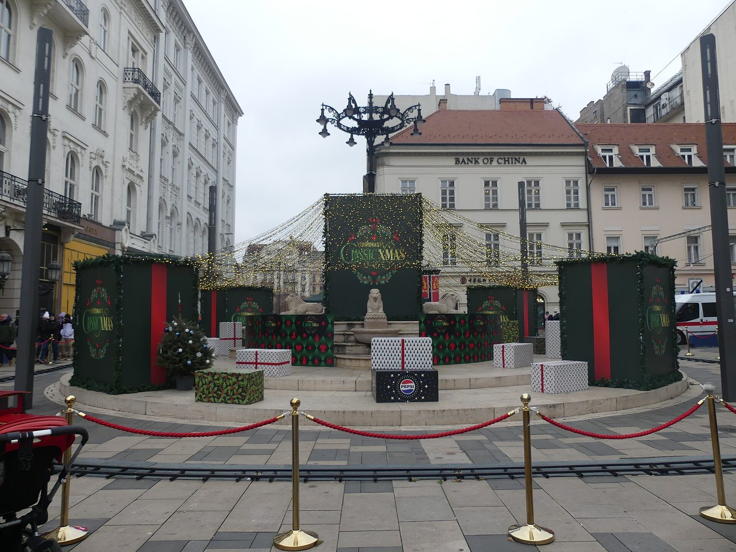 Vánočně vyzdobený podstavec sochy na Vörösmarty tér v Budapešti s kolejemi dětského vláčku a zlatými sloupky