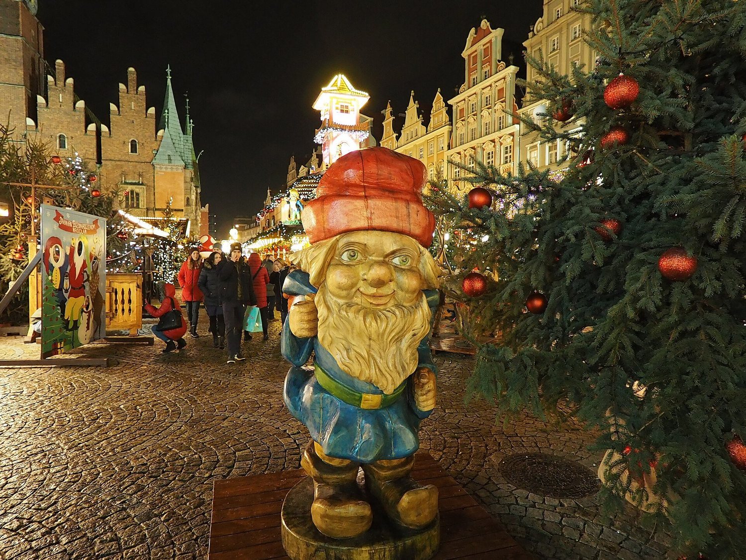 Dřevěná figurka trpaslíka s červenou čepicí mezi stánky vratislavského vánočního trhu večer