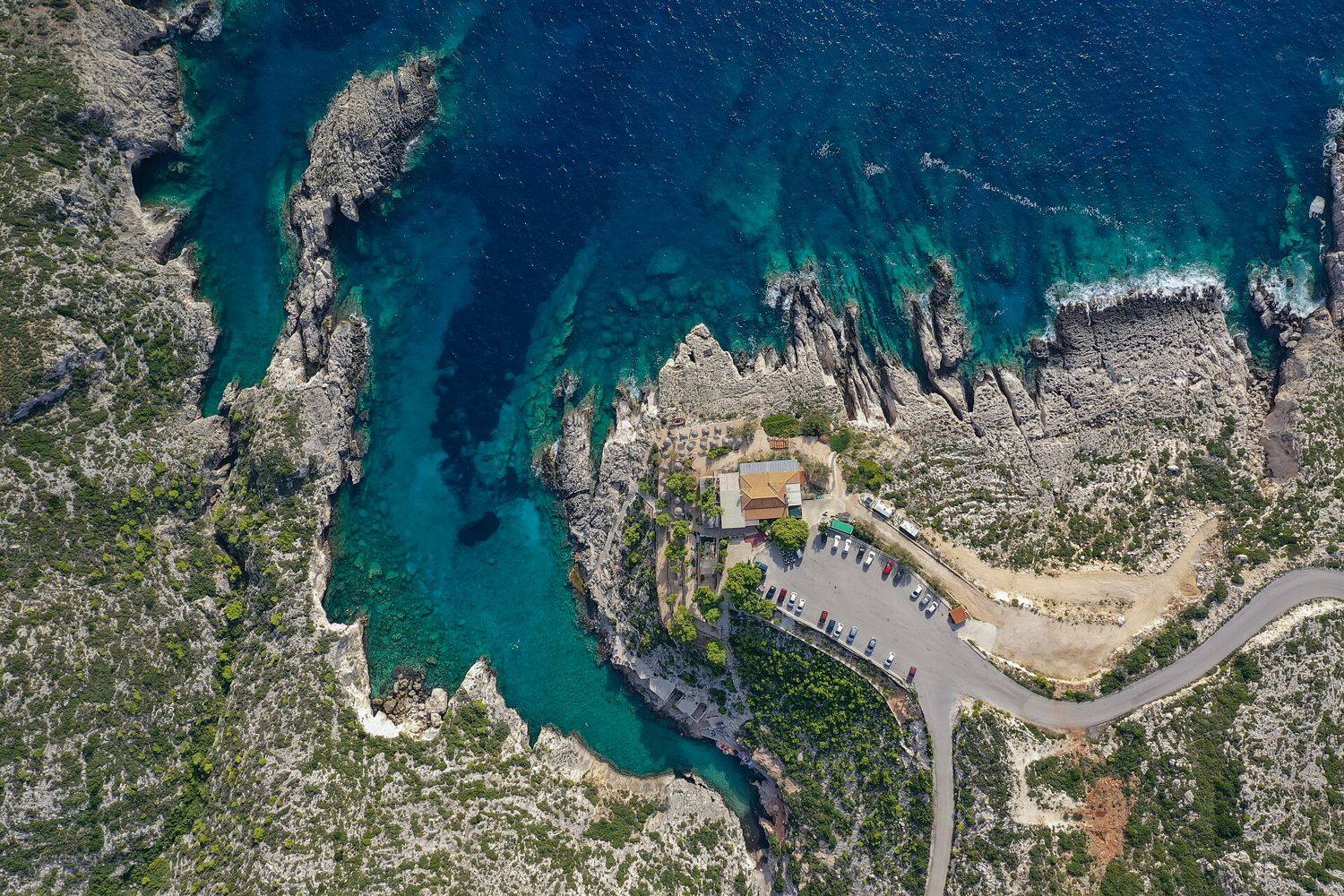 Zátoka Porto Limnionas shora: skalní plošiny, taverna, parkoviště a klikatá silnice